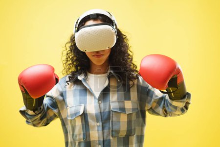 Téléchargez les photos : Portrait d'une jeune femme portant des lunettes VR et des gants de boxe tenant les mains en position de garde, jouissant d'un jeu de boxe, d'un joueur et d'un passe-temps sur fond jaune. - en image libre de droit