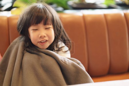 Photo for Portrait of  Asian adorable girl frowning looking toward the camera with copy space, while sitting on the sofa covering herself with an oversized daddy jacket, feeling  happy - Royalty Free Image