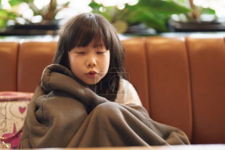 Téléchargez les photos : Portrait de fille adorable asiatique fronçant les sourcils vers l'appareil photo avec espace de copie, tout en étant assise sur le canapé se couvrant d'une veste papa surdimensionnée, se sentant heureuse - en image libre de droit