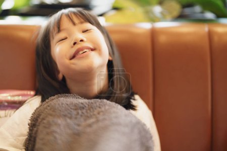 Téléchargez les photos : Portrait de fille adorable asiatique fronçant les sourcils vers l'appareil photo avec espace de copie, tout en s'asseyant sur le canapé se couvrant d'une veste papa surdimensionnée, se sentant ennuyé et pas heureux - en image libre de droit