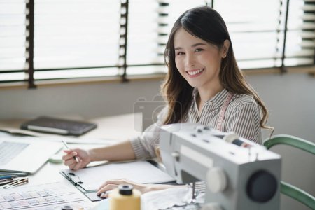 Photo for Smiling Asian Fashion Designer Crafting Trendy Clothing in a Boutique while looking at Camera. - Royalty Free Image