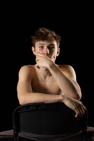 Photo for A portrait of 17 year old shirtless muscular teenage boy sititng on a chair against  a black background - Royalty Free Image