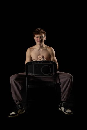 Photo for A portrait of 17 year old shirtless muscular teenage boy sititng on a chair against  a black background - Royalty Free Image