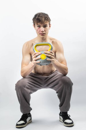 Photo for A 17 year old shirtless muscular teenage boy doing a squat with a kettlebell - Royalty Free Image