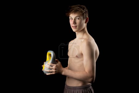 Photo for A 17 year old shirtless muscular teenage boy holding a kettlebell against a black background - Royalty Free Image