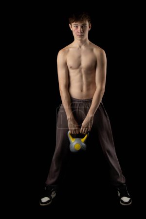 Photo for A 17 year old shirtless muscular teenage boy holding a kettlebell against a black background - Royalty Free Image