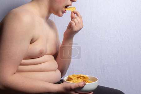 An overweight caucasian teenage boy eating a bowl of crisps
