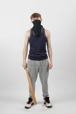 A Teenage Male Gang Member in a mask holding a baseball bat by his leg