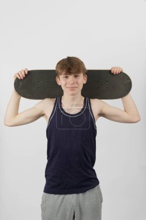 Ein fünfzehnjähriger Skateboard-Junge hält sich vor weißem Hintergrund ein Skateboard hinter den Kopf