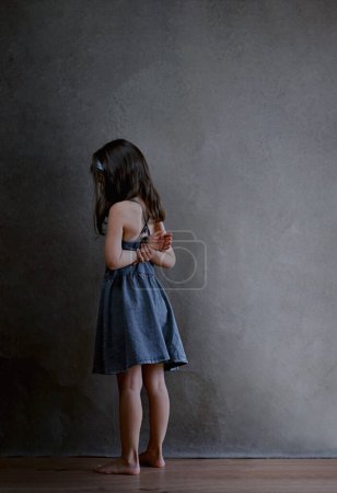 Bestraftes Mädchen steht in der Ecke und wird von der ganzen Welt beleidigt, Rückansicht eines Mädchens mit langen dunklen Haaren in einem blauen Sommerkleid, das die Hände auf dem Rücken vor dunklem Hintergrund hält. Hoch
