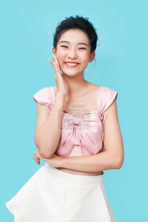 Photo for A beautiful asian young girl without acne or blemishes, white smile, touching face and looking happy, standing over cyan background - Royalty Free Image