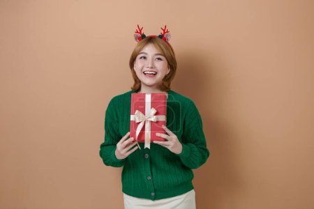 Photo for Cheerful young woman in a green sweater with deer antlers holds a Christmas gift and laughing - Royalty Free Image