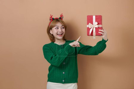 Photo for Smiling girl wearing deer headband and green knitted sweater is pointing with index finger at decorated gift box in her hand. - Royalty Free Image