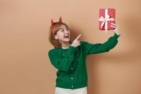 Photo for Smiling girl wearing deer headband and green knitted sweater is pointing with index finger at decorated gift box in her hand. - Royalty Free Image