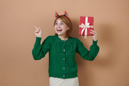Photo for Happy girl wearing Christmas deer horns holding a gift box, pointing to side at blank copy space - Royalty Free Image