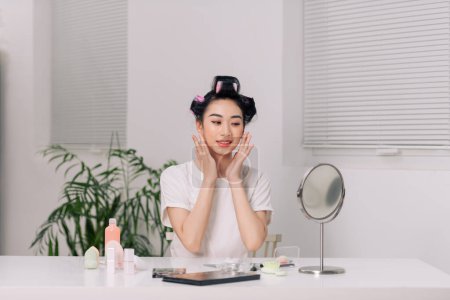 Foto de Bonita mujer asiática haciendo masaje lifting facial, sentada en la mesa en el dormitorio, mirando al espejo y sonriendo - Imagen libre de derechos