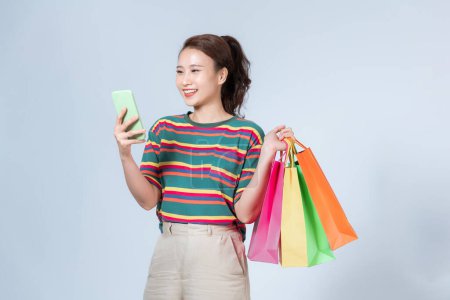 Foto de Hermosa mujer joven sosteniendo bolsas de la compra y el uso de su teléfono inteligente con sonrisa, mientras que de pie sobre fondo blanco - Imagen libre de derechos