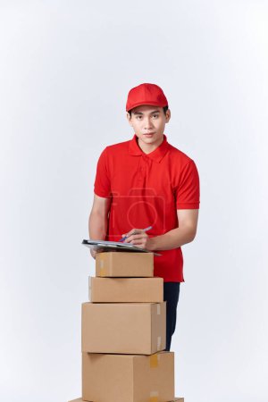Photo for Cheerful professional male mover in casual wear making notes, holding clipboard, standing near a stack of cardboard boxes while doing home relocation. Service concept - Royalty Free Image