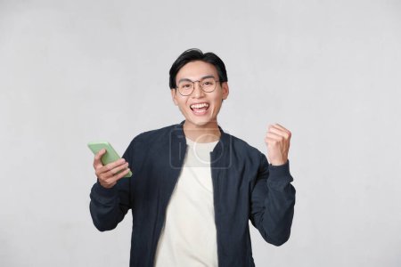 Photo for Excited young Asian man using smartphone and making winner gesture isolated - Royalty Free Image