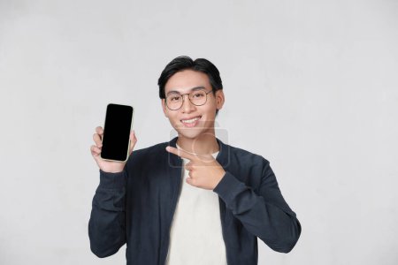 Foto de Cheerful asian guy pointing at smartphone with blank screen in his hand - Imagen libre de derechos