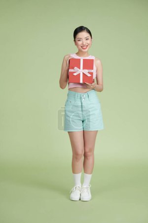 Photo for Happy woman holding gift box and looking at the camera while enjoys over green background - Royalty Free Image