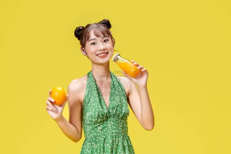 Téléchargez les photos : Excité asiatique femme boire boisson fraîcheur orange jus verre debout sur jaune fond - en image libre de droit