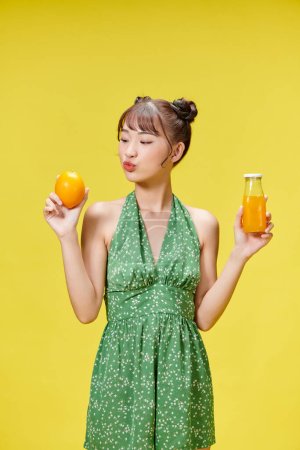 Foto de Chica positiva sosteniendo naranja fresca y jugo, fondo amarillo estudio - Imagen libre de derechos