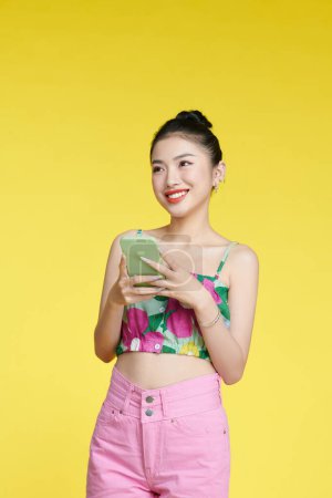 Téléchargez les photos : Plaisir jeune asiatique femme portant débardeur sur fond jaune en utilisant le téléphone - en image libre de droit