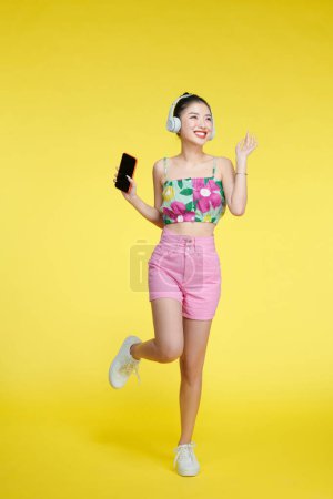 Téléchargez les photos : Heureuse femme asiatique joyeuse portant des écouteurs sans fil écoutant de la musique depuis un smartphone - en image libre de droit