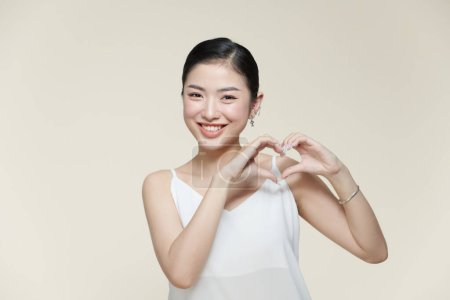 Photo for Portrait of positive young lady with healthy smooth skin holding hands in the shape of a heart near face - Royalty Free Image