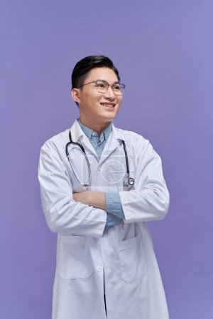 Foto de Sonriente joven médico varón con estetoscopio sobre el cuello en abrigo médico aislado en púrpura - Imagen libre de derechos