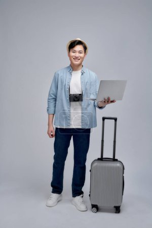 Téléchargez les photos : Jeune homme debout avec valise à bagages avec ordinateur portable concentré souriant joyeux - en image libre de droit
