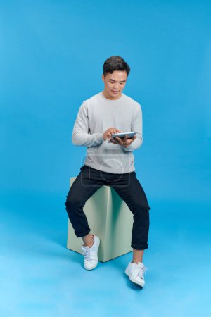 Téléchargez les photos : Joyeux jeune homme assis sur un cube géométrique et utilisant une tablette sur fond bleu - en image libre de droit