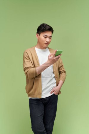 Téléchargez les photos : Image d'un homme asiatique heureux tenant son téléphone portable tout en étant isolé par un fond vert clair - en image libre de droit
