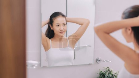 Téléchargez les photos : Femme en robe de chambre blanche attachant ses cheveux devant le miroir en photo de salle de bain lumineuse - en image libre de droit