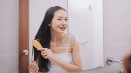 Téléchargez les photos : Joyeux attrayant jeune femme peignage cheveux mouillés avec peigne dans la salle de bain après la douche à la maison. Soins capillaires et cheveux sains - en image libre de droit