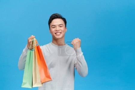 Téléchargez les photos : Jeune homme asiatique super heureux tenant des sacs à provisions, isolé sur fond bleu. - en image libre de droit