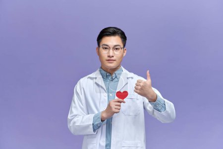 Téléchargez les photos : Jeune homme médecin tenant coeur en papier, pouce levé avec les doigts sur fond violet - en image libre de droit