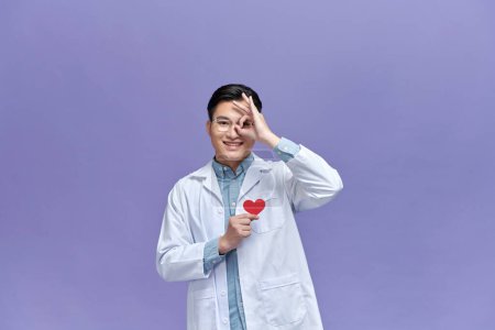 Téléchargez les photos : Médecin homme tenant coeur de papier rouge avec le visage heureux souriant faire ok signe avec la main sur l'œil regardant à travers les doigts - en image libre de droit