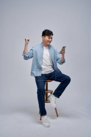 Photo for Young Asian man play game on mobile phone application and raising his arm up with celebrating success - Royalty Free Image