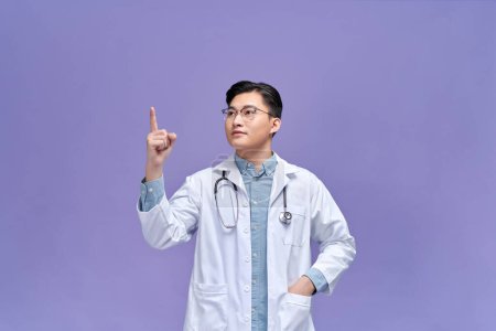 Téléchargez les photos : Jeune homme portant l'uniforme de médecin avec un sourire du visage pointant avec la main et le doigt sur le côté - en image libre de droit