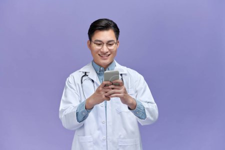 Foto de Retrato del médico masculino usando teléfono móvil y sonriendo aislado sobre fondo púrpura. - Imagen libre de derechos