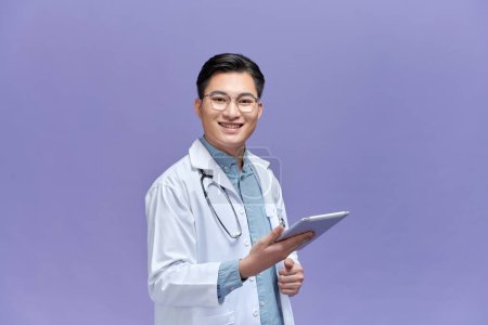 Foto de Sonriente joven médico de pelaje médico usando tableta digital aislada en púrpura - Imagen libre de derechos