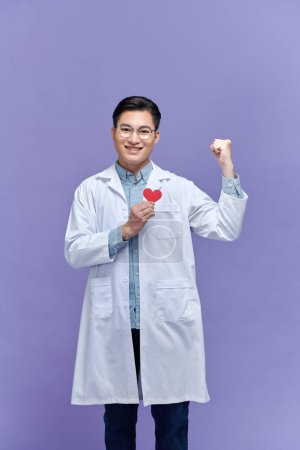 Foto de Médico hombre sosteniendo el corazón rojo en la clínica gritando orgulloso con el brazo levantado - Imagen libre de derechos