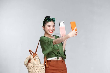 Foto de Una mujer sonriente tomando una selfie mientras sostiene un pasaporte con billete de avión sobre fondo blanco - Imagen libre de derechos
