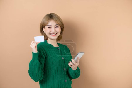 Foto de Retrato de mujer sonriente pagando con tarjeta de crédito de plástico en la aplicación de teléfono inteligente, de pie con teléfono móvil y tarjeta bancaria sobre fondo beige - Imagen libre de derechos