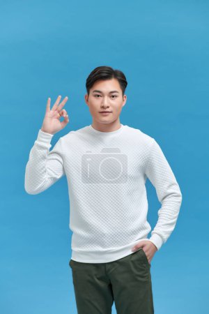 Téléchargez les photos : Jeune homme asiatique montrant ok signe de la main et souriant tout en se tenant sur fond bleu - en image libre de droit
