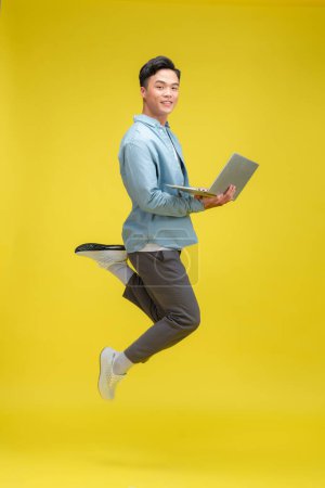 Foto de Foto de un joven excitado saltando aislado sobre un fondo de pared amarillo usando una computadora portátil
. - Imagen libre de derechos