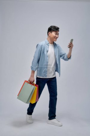 Foto de Joven asiático hombre con bolsas de compras está utilizando un teléfono móvil y sonriendo mientras hace compras - Imagen libre de derechos