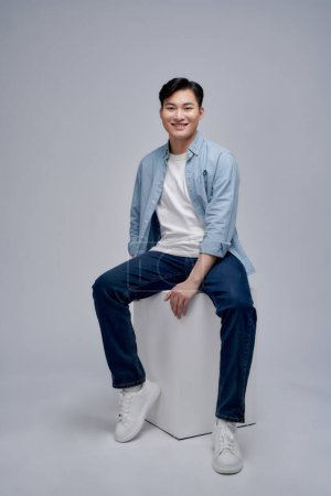 Photo for Young asian man posing while seated on box looking away from the camera in studio background - Royalty Free Image